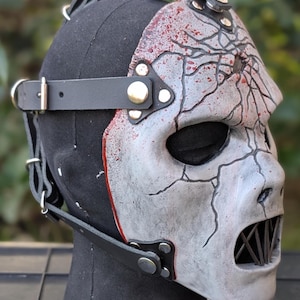 May include: A distressed, cracked mask with a gray and white finish, featuring black lines and red accents. The mask has a menacing open mouth design and is secured with black leather straps and metal hardware. The mask is displayed on a black mannequin head.