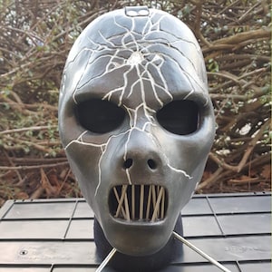 May include: A dark gray mask with a cracked design and a wide open mouth with vertical bars. The mask has a smooth finish and is likely made of a hard material. The cracks are highlighted with white lines.