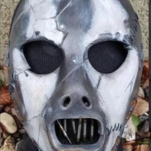 May include: A distressed, metallic-looking mask with a cracked, weathered appearance. The mask features large, oval eye openings covered with black mesh, a gaping mouth revealing vertical bars, and stitched details. The color scheme is primarily silver and black.