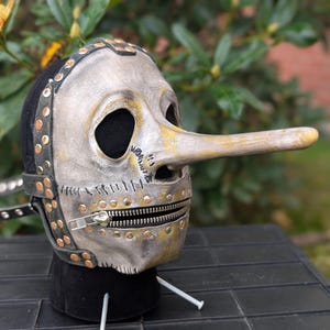 May include: A distressed silver mask with a long, yellowed nose and a zipper mouth. The mask has black eye holes and is adorned with rivets and leather straps. The mask is likely for costume or theatrical use.