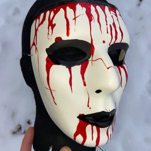 May include: A white mask with black eye sockets and lips, and red paint dripping down the face. The mask is mounted on a black head form. The mask is likely for a costume or theatrical performance.