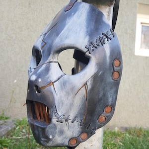 May include: A handcrafted, gray and silver mask with a stitched design and visible cracks. The mask has a menacing appearance with a wide open mouth revealing wooden slats. The mask is held in place by a black strap.