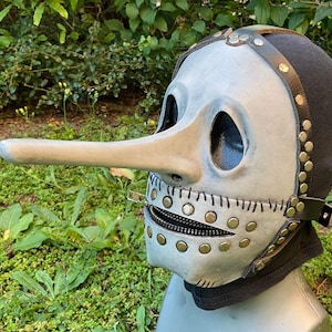 May include: A gray plague doctor mask with a long, curved nose and large eye openings. The mask features a zipper mouth, brass rivets, and leather straps, set against a black hood. The mask is a unique costume accessory.