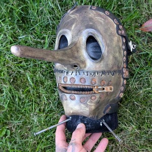 May include: A metallic mask with a long nose and a zipper mouth. The mask has a distressed, aged appearance with rivets and stitching details. The eye holes are large and dark. The mask is resting on a black base, held by a hand.
