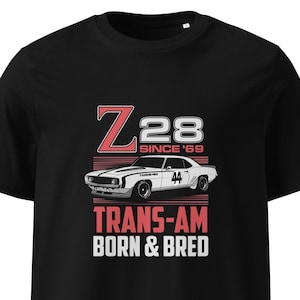Puede incluir: Camiseta negra con un diseño gráfico. El diseño incluye el texto "Z28 SINCE '69" en rojo y blanco, y una ilustración de un coche blanco con el número "44". Debajo del coche están las palabras "TRANS-AM BORN & BRED".