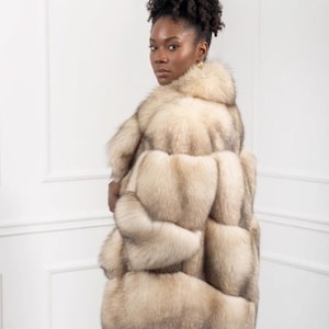 May include: A light-coloured fur coat with a layered design. The coat features a high collar and a textured appearance, with shades of cream and grey. The model is wearing gold earrings and has dark, curly hair.