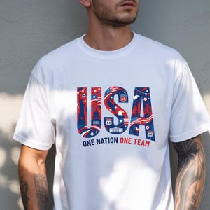 May include: A white short-sleeved t-shirt featuring the text USA in large blue letters filled with red and white patriotic graphics including the Statue of Liberty, Route 66 signs, and eagles. Below the text is the phrase ONE NATION ONE TEAM.