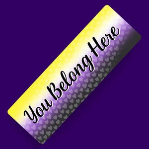 Puede incluir: Una pegatina rectangular con un fondo degradado que transita de amarillo a blanco y a morado, superpuesto con un patrón repetitivo de pequeños corazones grises. El texto "You Belong Here" está impreso en una fuente cursiva negra.