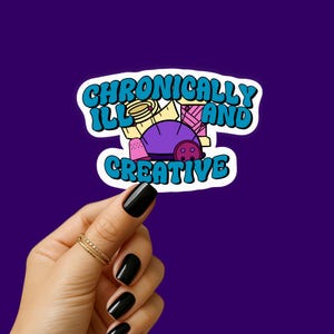 Op de afbeelding: Een sticker met de tekst "Chronically Ill and Creative" in een blauw en wit bubbellettertype. De sticker bevat illustraties van naaiartikelen, waaronder een paars speldenkussen, klosjes garen en een vingerhoed, op een paarse achtergrond.