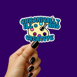 May include: A sticker with the text "Chronically Ill and Creative" in blue and white, featuring a yellow artist's palette with colorful paint splotches and a paintbrush. The sticker is held by a hand with black nail polish and gold rings, set against a purple background.
