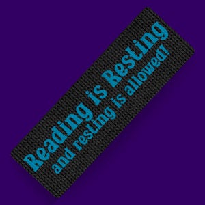 Può includere: Segnalibro rettangolare nero con texture a maglia e testo blu "Reading is Resting and resting is allowed!". Sfondo viola, perfetto per la lettura.