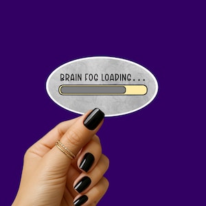Pode incluir: Um adesivo oval com o texto "BRAIN FOG LOADING..." e uma barra de progresso, em um fundo roxo. A barra de progresso está parcialmente preenchida. A mão que segura o adesivo tem esmalte preto e anéis dourados.