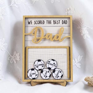 May include: Wooden soccer-themed frame with the text "WE SCORED THE BEST DAD" and the word "Dad" in cursive. The frame holds five soccer ball magnets with player names and numbers. The frame is supported by a wooden stand.