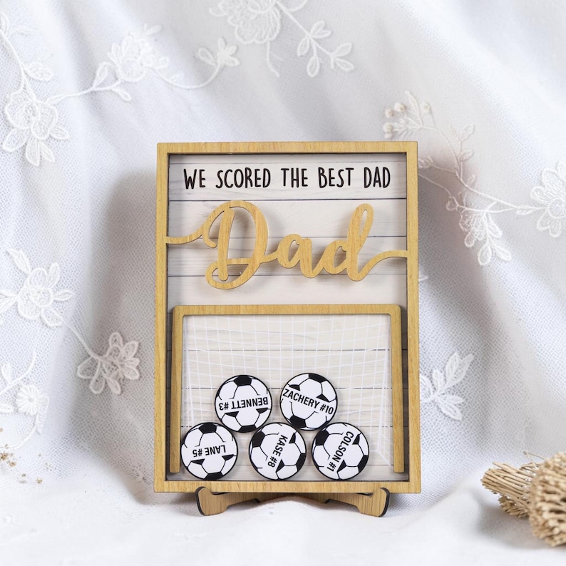 May include: Wooden soccer-themed frame with the text "WE SCORED THE BEST DAD" and the word "Dad" in cursive. The frame holds five soccer ball magnets with player names and numbers. The frame is supported by a wooden stand.