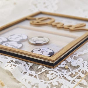 May include: A wooden shadow box with the word "Dad" cut out at the top. Inside are several soccer ball-themed tokens with names and numbers. The frame has a black stripe and a silver accent.