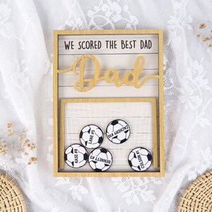 May include: A wooden framed sign with the text "WE SCORED THE BEST DAD" and a cutout word "Dad". Below, five soccer ball magnets are arranged in front of a net. Each ball has a name and number.