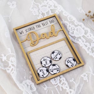 May include: A wooden framed keepsake with the words "WE SCORED THE BEST DAD" and "Dad" in a cursive font. The frame holds five soccer ball-shaped tokens with names and numbers. The frame is 20 cm wide and 15 cm tall.