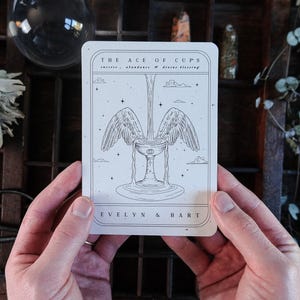 Pode incluir: Uma carta de tarot com uma ilustração detalhada de um cálice alado, com o texto "THE ACE OF CUPS" e "EVELYN & BART". A carta é segurada em duas mãos, sobre um fundo de madeira rústica.