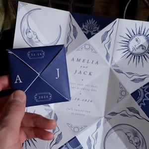 May include: Navy blue and white wedding invitation with celestial illustrations. The invitation features a folded design with a moon, sun, and floral motifs. The invitation includes the names "Amelia and Jack" and the date "10.10.2026".