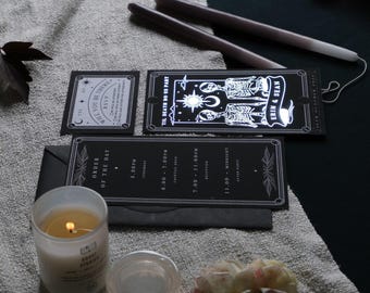 May include: A gothic-themed wedding invitation suite featuring black cards with white text and illustrations. The suite includes an RSVP card, an order of the day card, and a card with skeletons. A lit candle and taper candles are also present.