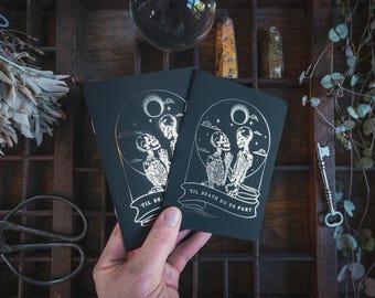 May include: Two black cards with silver foil illustrations of skeletons under a moon and clouds, with the text "TIL DEATH DO US PART". The cards are held in a hand, with a dark wooden background.