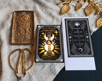 Op de afbeelding: Een assortiment trouwbriefpapier en decoratieve items. Inclusief een messing dienblad met een zon- en maanontwerp, een gouden schaar en twee uitnodigingskaarten met hemelse afbeeldingen. Eén kaart leest "Jessica & Ashley" en "Save the Date".