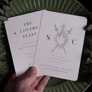 May include: Two light pink wedding menu cards with black text. One card reads "The Lovers Feast" and lists menu items. The other card has a heart pierced by swords and the initials "S" and "C". The text reads "Suzy & Carina, Tuesday 18th March 2025, Bolton Castle".