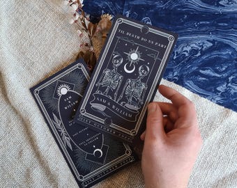 May include: Dark navy wedding invitations with white illustrations of skeletons, suns, and moons. The text reads "Til Death Do Us Part" and "Sam & William", with the date "31st October 2022". The invitations are rectangular with a decorative border.