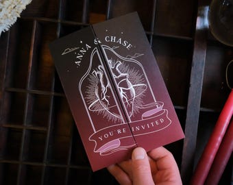 May include: A folded invitation card with a dark red and black gradient background. The card features a white illustration of a heart and the text "ANNA & CHASE" and "YOU'RE INVITED".