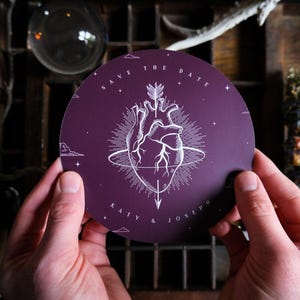 May include: A round, dark purple "Save the Date" card featuring a white anatomical heart design with an arrow. The card includes the names "Katy & Joseph" and is held by two hands. The background includes a wooden shelf and other decorative items.