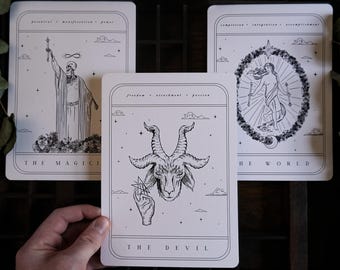 May include: Three tarot cards with black and white illustrations. The cards feature images of a skeleton, a goat head, and a figure within a circular frame. Text includes "The Magician", "The Devil", and "The World".