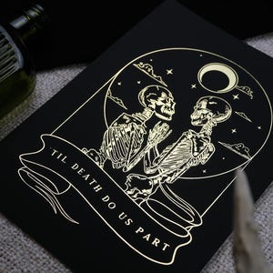 May include: Black art print with a gold illustration of two skeletons embracing, set within a circle with a moon and stars. The words "Til Death Do Us Part" are on a banner. The artwork has a gothic theme.