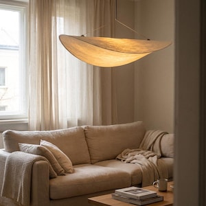 Puede incluir: Un salón moderno en tonos beige, con una gran lámpara colgante curva. La luz cuelga sobre un sofá beige con cojines y una manta. Unas cortinas transparentes enmarcan una ventana y una mesa de centro de madera está frente al sofá.