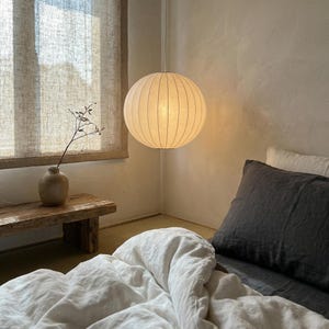 May include: A spherical, cream-coloured pendant light with vertical lines hangs in a bedroom. The light is suspended from a white cord. A bed with white linens and a dark grey pillow is in the foreground. A wooden bench and vase are visible.