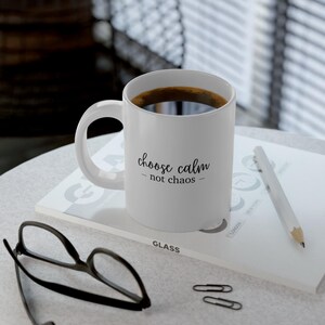 Puede incluir: Taza de cerámica blanca llena de café oscuro, con el texto "choose calm - not chaos" en escritura negra. La taza está sobre una superficie blanca con un lápiz, gafas y clips.