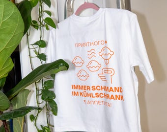 Puede incluir: Camiseta blanca con gráfico naranja de alimentos y texto en varios idiomas. La camiseta está colgada de una percha rosa en una escalera de metal. Una planta verde está en primer plano.