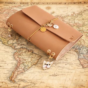 May include: A tan leather journal with a flap closure, gold-coloured eyelets, and a gold elastic band. The journal is adorned with charms, including a bulldog, a heart, and initial charms. The journal rests on a vintage world map.