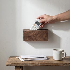 Könnte beinhalten: Ein rechteckiger, an der Wand befestigter Holzorganizer mit einer Fernbedienung. Der Organizer ist aus dunklem Holz gefertigt und an einer weißen Wand befestigt. Eine Hand greift nach der Fernbedienung. Ein Stapel Post und eine Tasse stehen auf einem Holztisch.