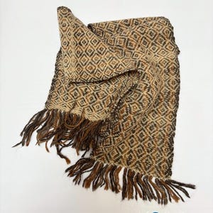 May include: A woven scarf featuring a repeating diamond pattern in shades of brown, tan, and cream. The rectangular accessory has long, loose fringe along the short edges. The bottom right corner displays the text KOSHA Argentina with a blue icon.