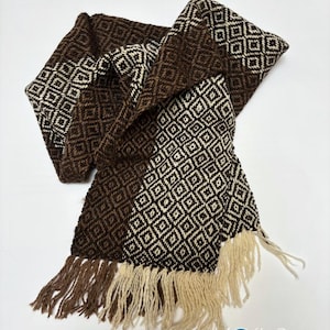 May include: A woven scarf featuring a repeating geometric diamond pattern in dark brown and cream colors. The scarf has a textured appearance and is finished with long, loose fringe tassels at the ends in matching dark brown and cream yarn colors.