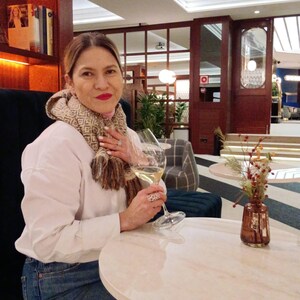 May include: A white long-sleeved shirt, blue denim jeans, and a beige and brown patterned scarf. The outfit includes a large silver ring with a circular cutout design on the finger and a clear glass of white wine held in the hand.