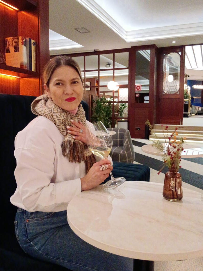 May include: A white long-sleeved shirt, blue denim jeans, and a beige and brown patterned scarf. The outfit includes a large silver ring with a circular cutout design on the finger and a clear glass of white wine held in the hand.