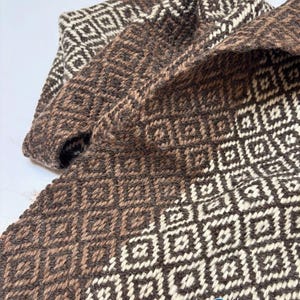 May include: A woven textile featuring a repeating geometric square pattern in shades of dark brown and cream. The fabric displays a textured, interlaced appearance with a consistent grid design throughout the material, including the logo KOSHA.