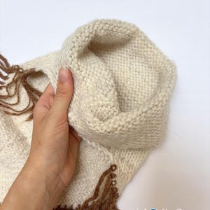 May include: A cream-coloured knitted cowl or scarf featuring a textured stitch pattern and long, brown fringe detailing along the edges. The item includes a logo in the bottom right corner that reads Kosha Argentina with a blue circular icon.