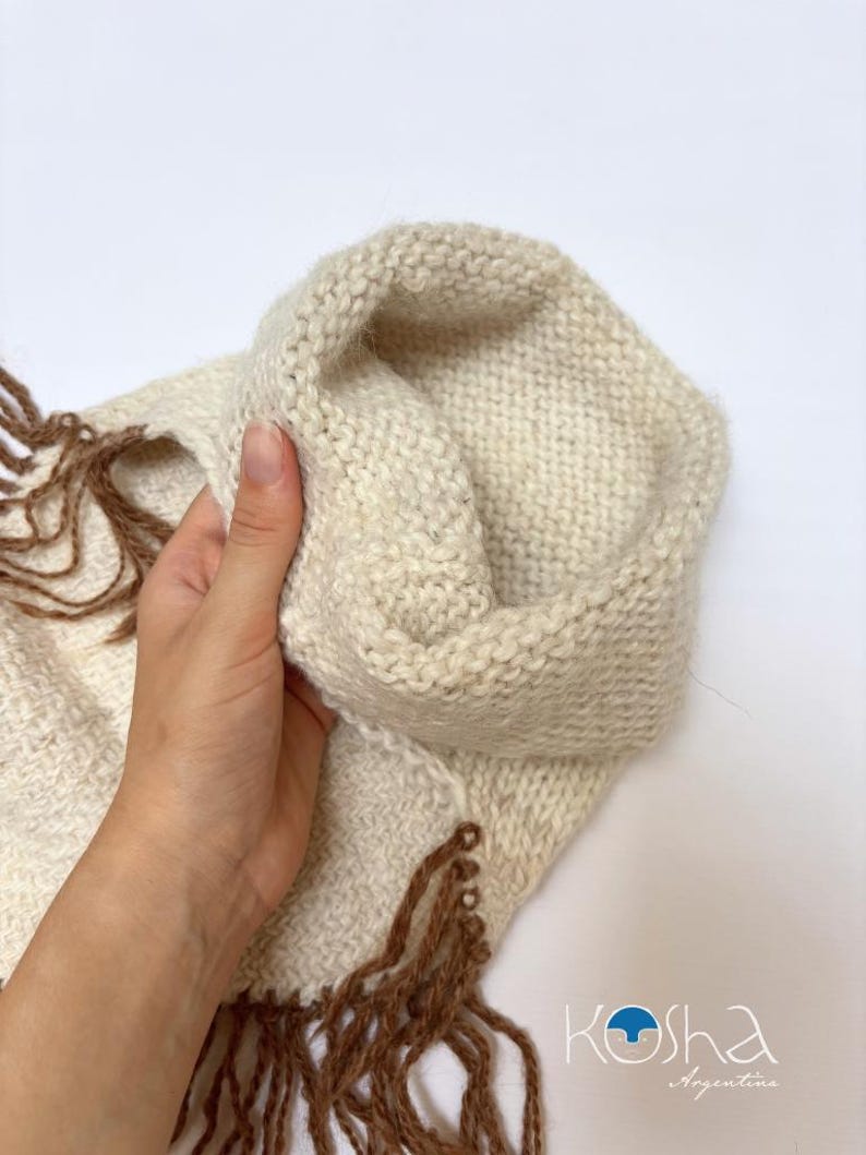 May include: A cream-coloured knitted cowl or scarf featuring a textured stitch pattern and long, brown fringe detailing along the edges. The item includes a logo in the bottom right corner that reads Kosha Argentina with a blue circular icon.