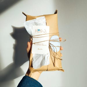 Puede incluir: Un paquete rectangular envuelto en papel marr&oacute;n y atado con cordel fino. Una tarjeta blanca con el texto Kosha y texto impreso adicional est&aacute; metida bajo el cordel, acompa&ntilde;ada de una peque&ntilde;a etiqueta de papel circular de color azul claro.