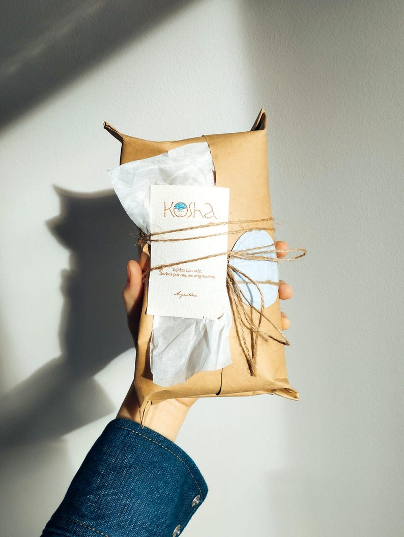 Puede incluir: Un paquete rectangular envuelto en papel marr&oacute;n y atado con cordel fino. Una tarjeta blanca con el texto Kosha y texto impreso adicional est&aacute; metida bajo el cordel, acompa&ntilde;ada de una peque&ntilde;a etiqueta de papel circular de color azul claro.