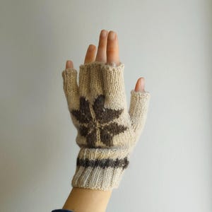 May include: A beige fingerless glove featuring a dark brown star-shaped pattern on the back of the hand and a ribbed cuff. The knit accessory has individual openings for the fingers and thumb, showcasing a textured yarn construction throughout.