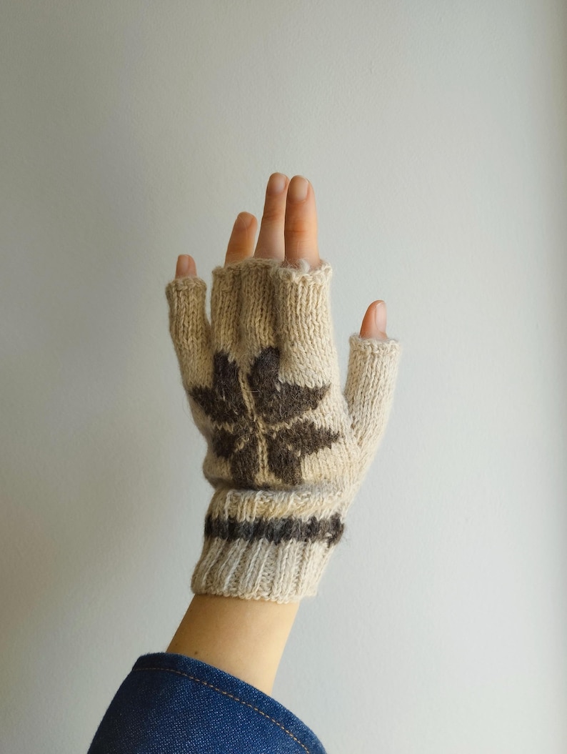 May include: A beige fingerless glove featuring a dark brown star-shaped pattern on the back of the hand and a ribbed cuff. The knit accessory has individual openings for the fingers and thumb, showcasing a textured yarn construction throughout.