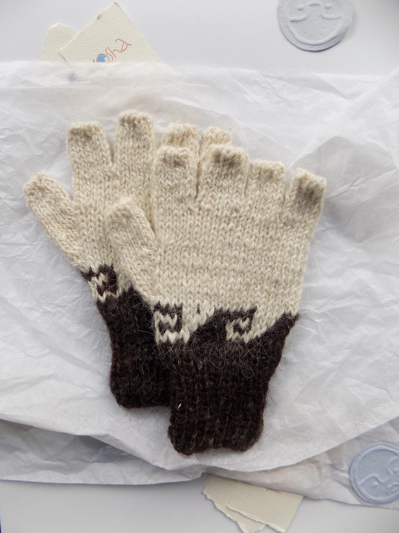 May include: A pair of fingerless gloves featuring a two-tone design with a cream-colored upper section and a dark brown ribbed cuff. The transition between the colors displays a geometric pattern, and the knit texture is visible throughout.